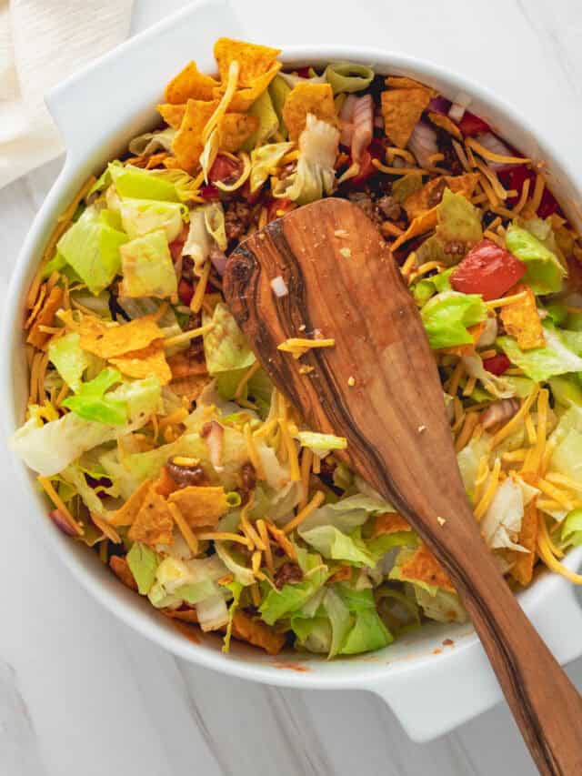 A serving spoon resting on top of a Dorito taco salad in a white casserole dish.