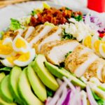 Chicken Cobb salad sitting on a bed of romaine lettuce and topped with jammy eggs, feta cheese, chopped bacon, and tomatoes.