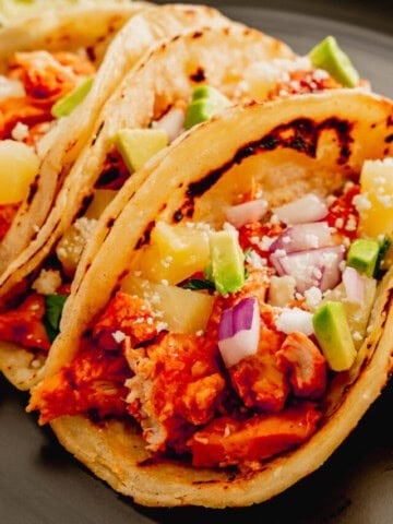 Instant Pot Chicken Al Pastor in corn tortillas topped with cojita cheese, pineapple chunks, and red onion.