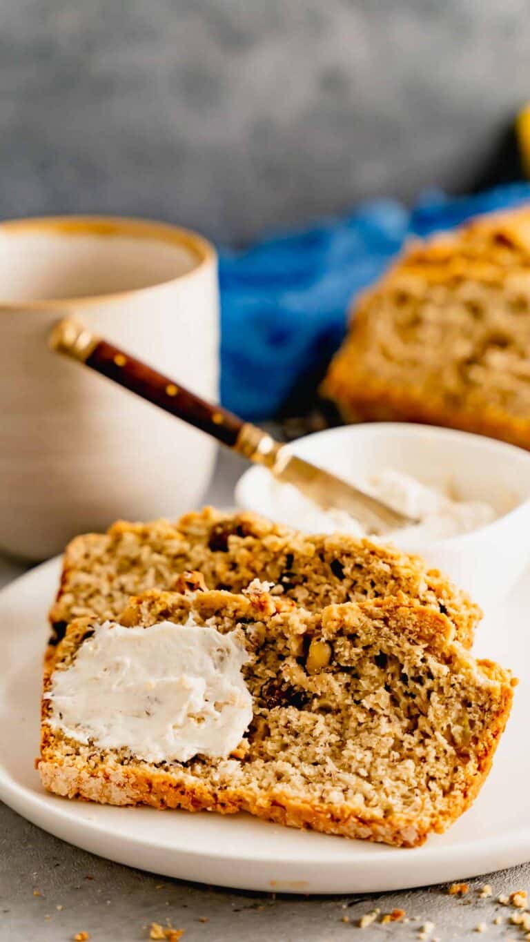 A white plate with two slices of banana nut bread with a coffee mug and cream cheese.