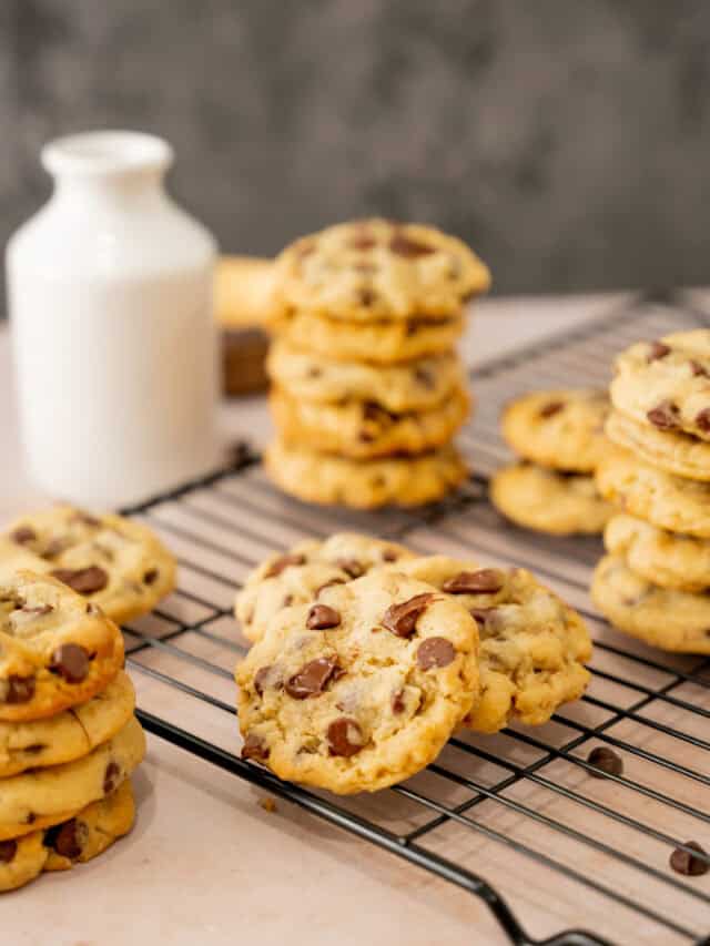 Scattered chocolate chip cookies on a cooling rack with a small jar of milk in the background.