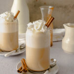 Two glasses of chai tea latte with whipped cream and cinnamon sticks.