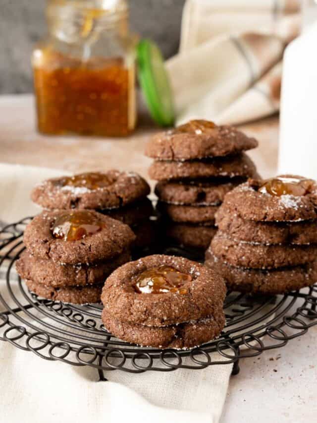 A square fig preserves jar in the background of a plate of chocolate thumbprint cookies.