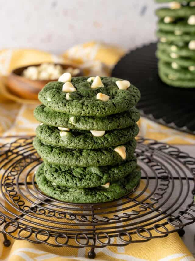 A stack of green velvet cookies with white chocolate chips on a wire round rack with a yellow background.
