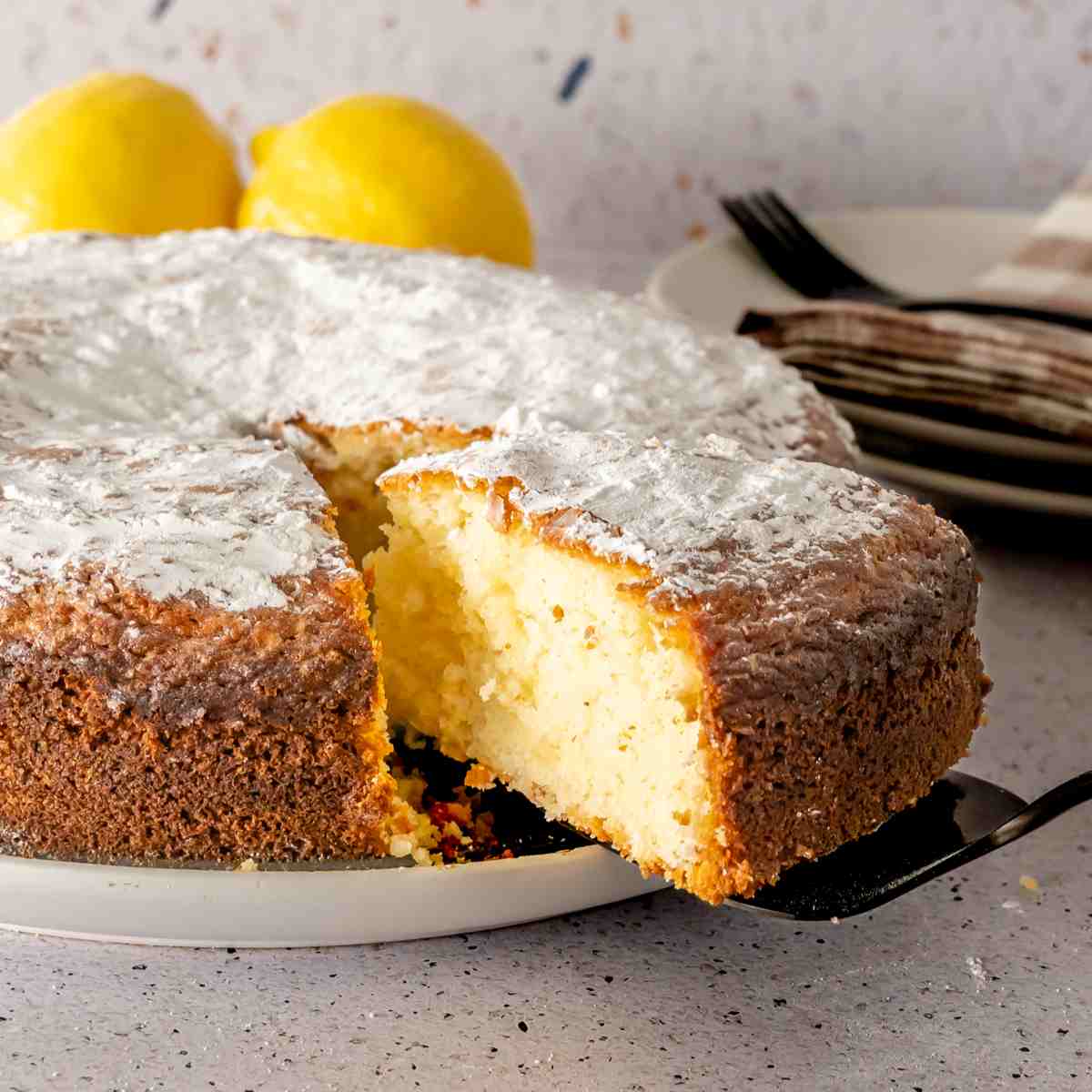 A black cake server pulling a slice of powdered sugar topped lemon and yogurt cake.