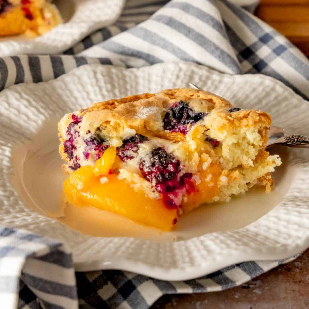 A slice of blueberry peach cobbler cake on a decorative plate with a black fork.