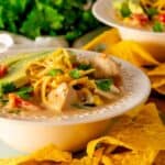 A rimmed bowl with chicken taco soup garnished with cheese, avocado slices, and tortilla chips.