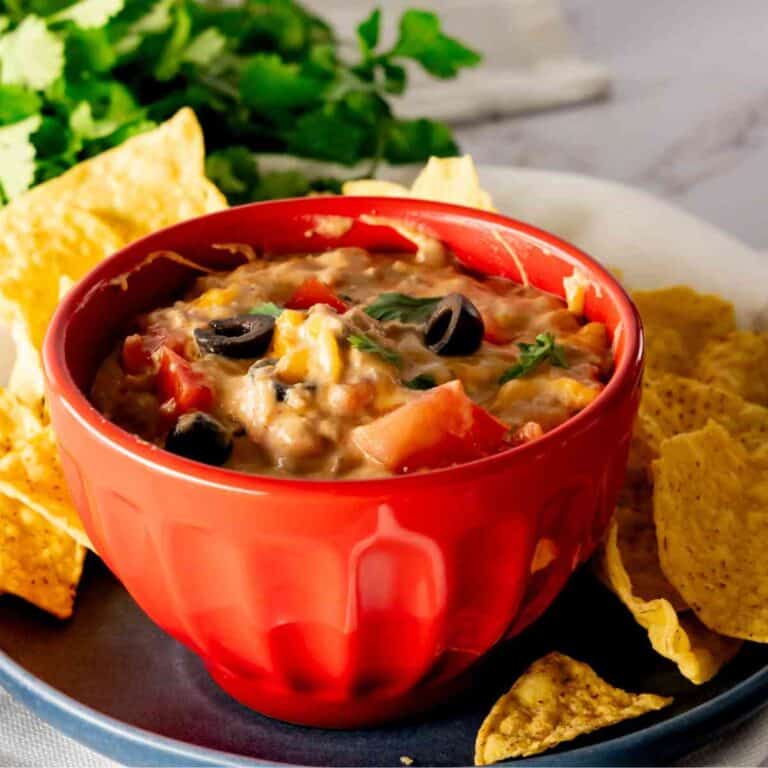 A bowl of taco dip with toppings with a slow cooker in the background.