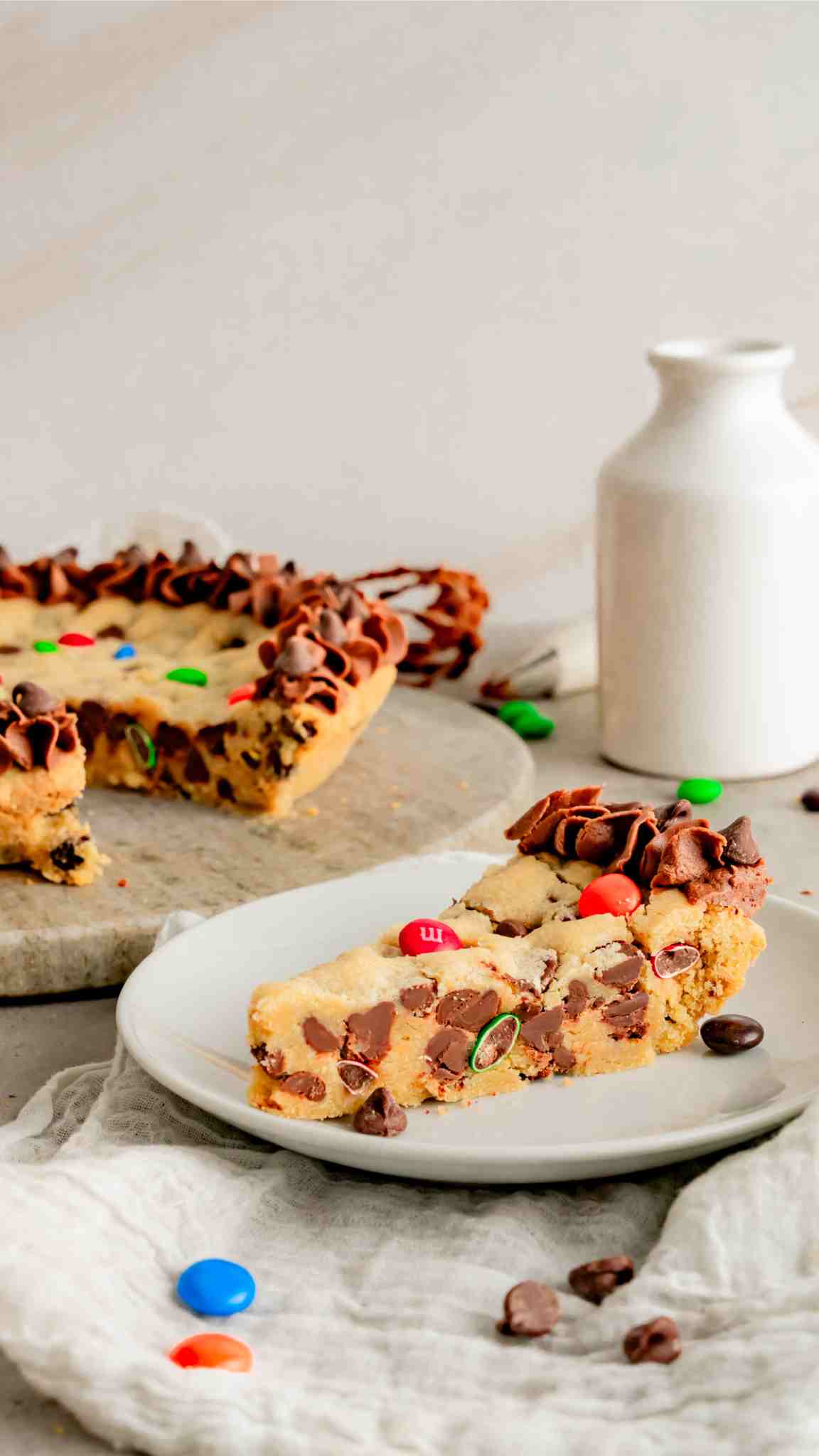 A slice of cookie cake with chocolate buttercream on a plate with a ceramic cup of milk.
