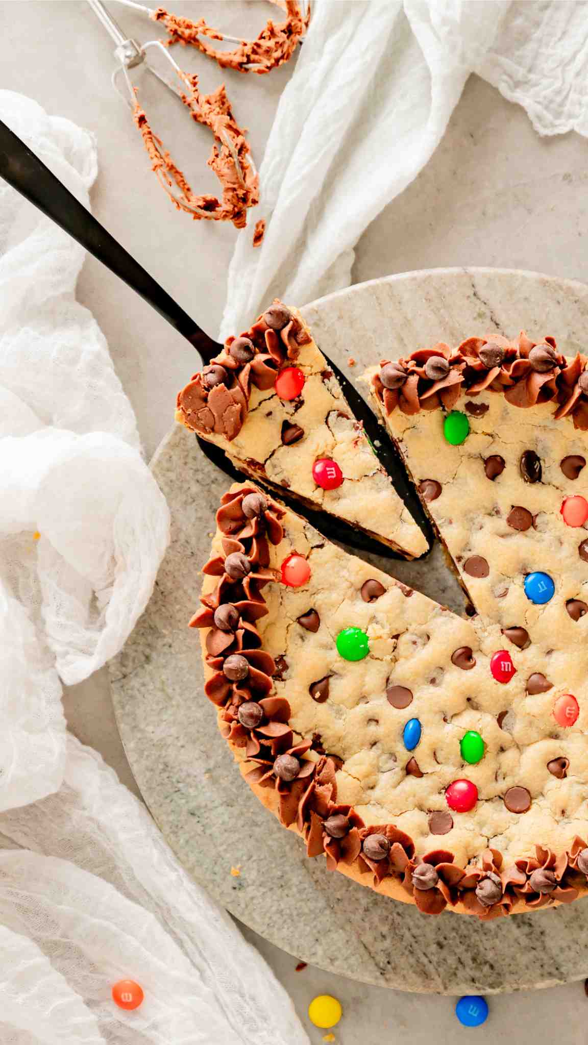 Overhead view of a cookie cake with a slice being taken out of it topped with chocolate chips and mms.