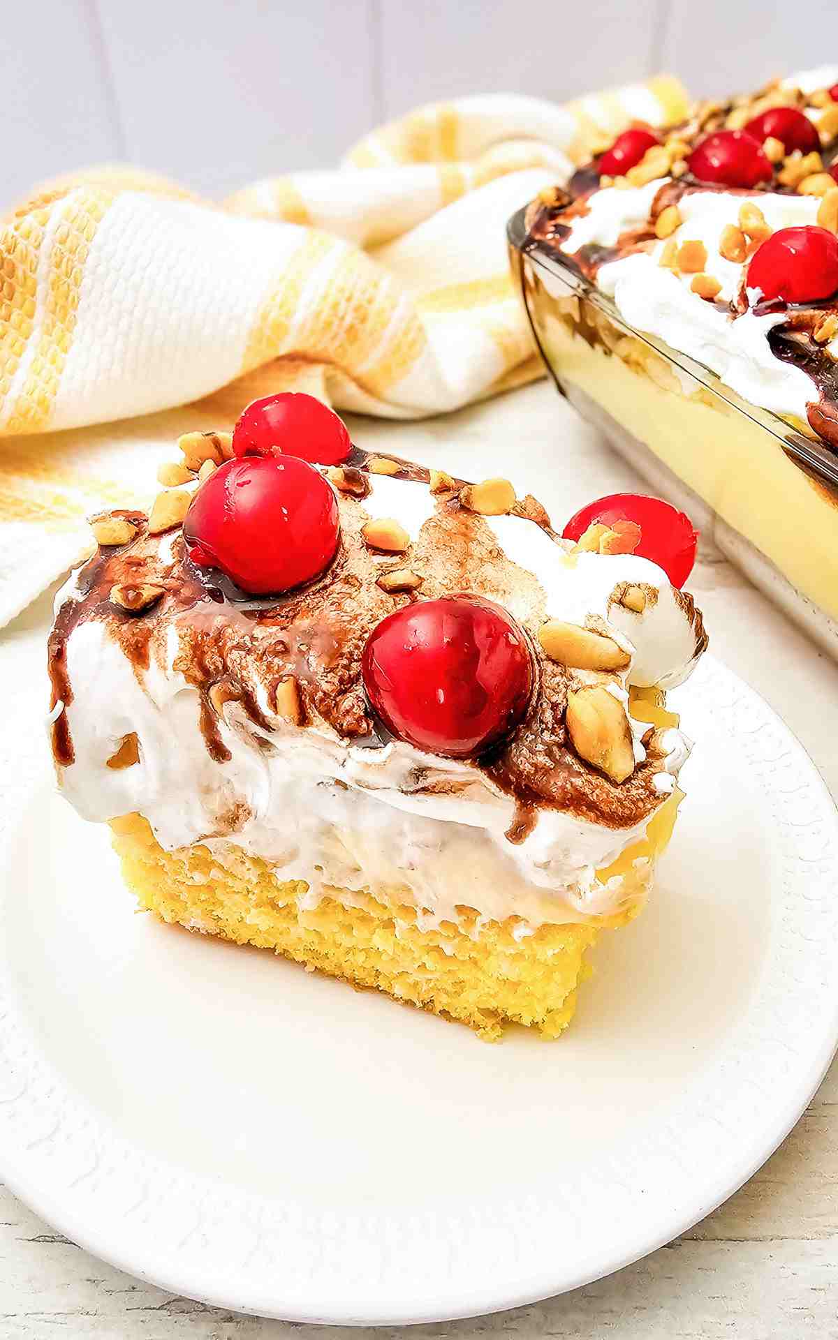 A slice of banana split cake on a plate topped with chocolate syrup and cherries.