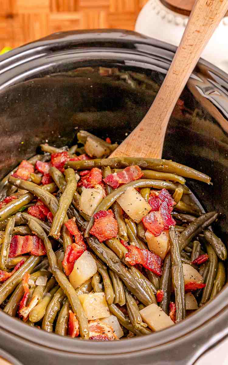 A wooden spoon in a slow cooker filled with southern green beans with cooked bacon.